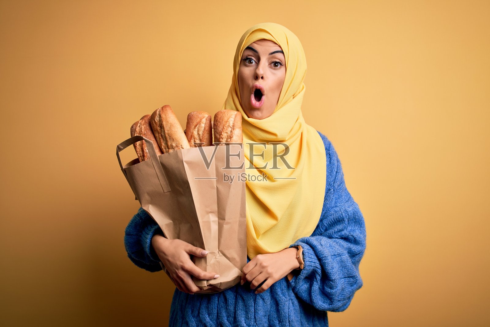 年轻漂亮的黑发阿拉伯妇女戴着伊斯兰的头巾,拿着纸袋和面包吓了一跳,脸上带着惊讶、害怕和兴奋的恐惧表情照片摄影图片