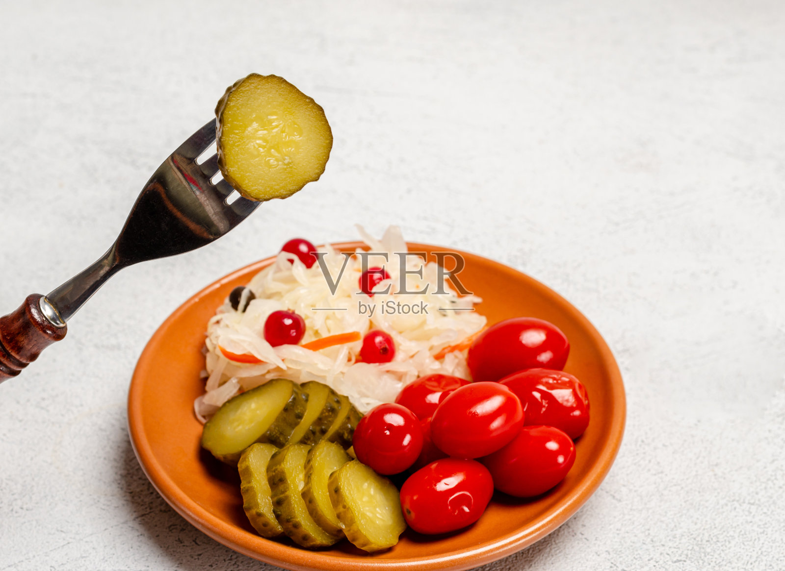 自制发酵制品放在盘子上,有酸菜、腌番茄、泡菜,背景浅灰色。叉子上放着一片腌黄瓜。照片摄影图片