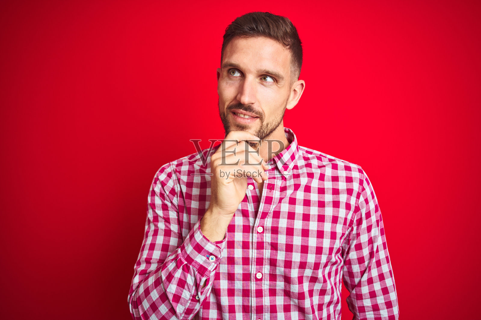 红底沉思的年轻英俊男子,手托下巴,面带微笑,似在思考问题照片摄影图片