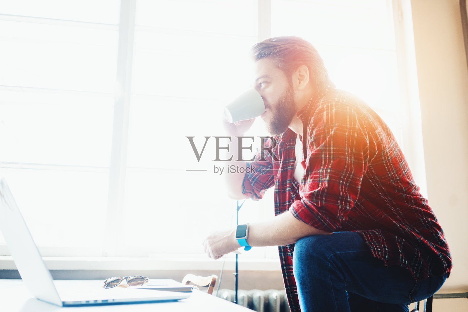 年轻的络腮胡男子在明亮的大办公室里用笔记本电脑工作,在阳光明媚的窗前喝咖啡照片摄影图片