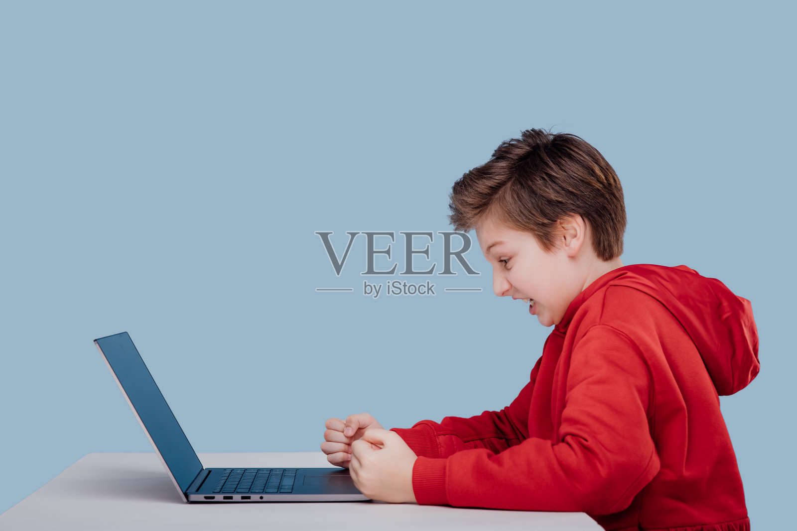 侧视图上瘾的愤怒的孩子男孩在红色运动衫在桌子上玩游戏在工作室的蓝色背景照片摄影图片