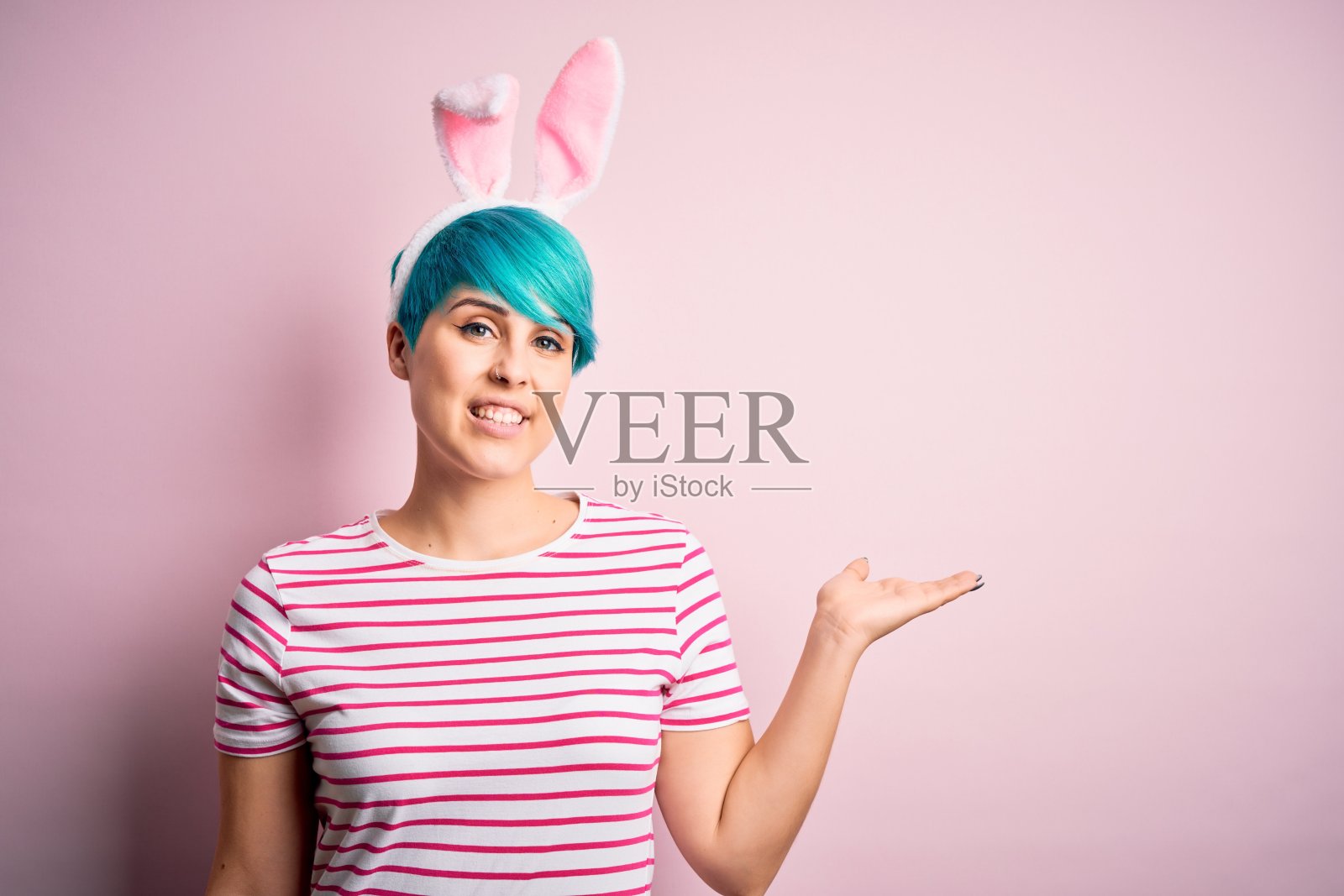 年轻女子,有时尚的蓝色头发,戴着复活节兔子耳朵,粉红色的背景,微笑着,用手掌看着镜头。照片摄影图片