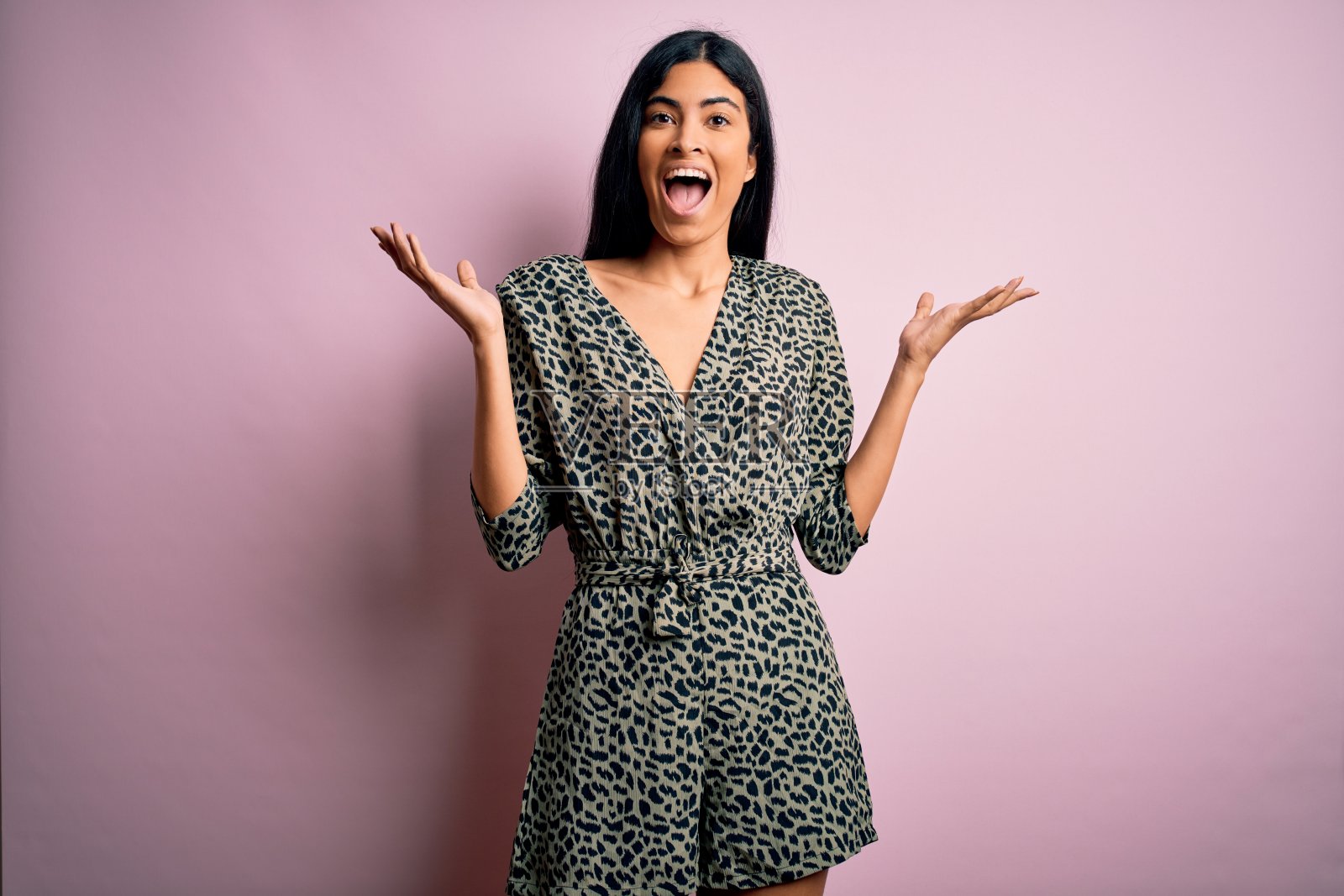 年轻美丽的西班牙时尚女人穿着休闲连衣裙在粉红色的背景庆祝疯狂和惊讶的成功,举起手臂,睁开眼睛兴奋地尖叫。赢家的概念照片摄影图片