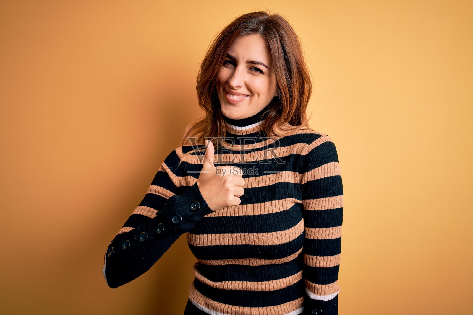 年轻漂亮的棕发女子穿着条纹高领毛衣,在黄色背景前,用手做出快乐的竖起大拇指的手势。表示赞同,看着镜头,展示成功。照片摄影图片