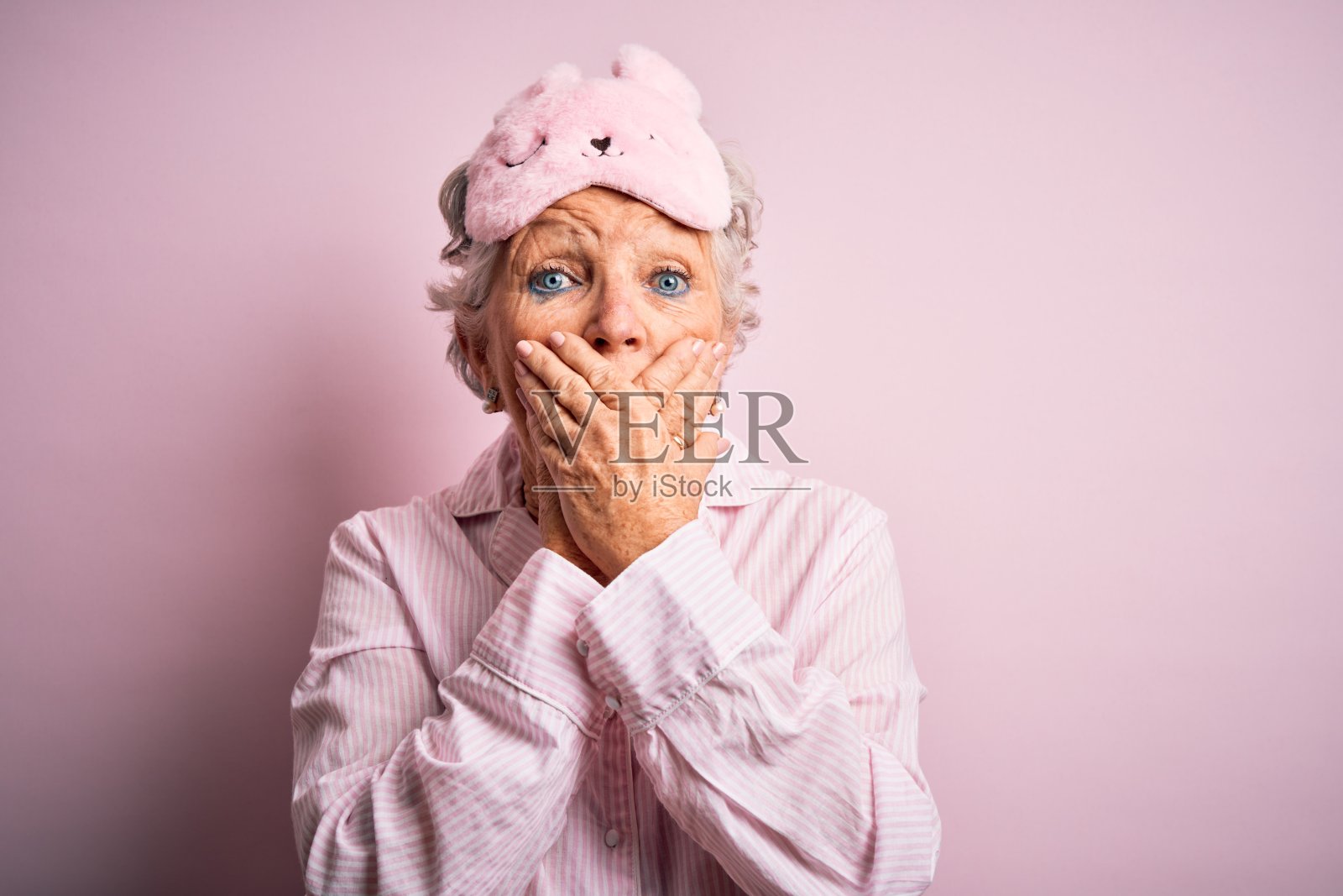 高级美女戴着睡眠面罩和睡衣在孤立的粉红色背景震惊掩盖嘴与手的错误。秘密的概念。照片摄影图片