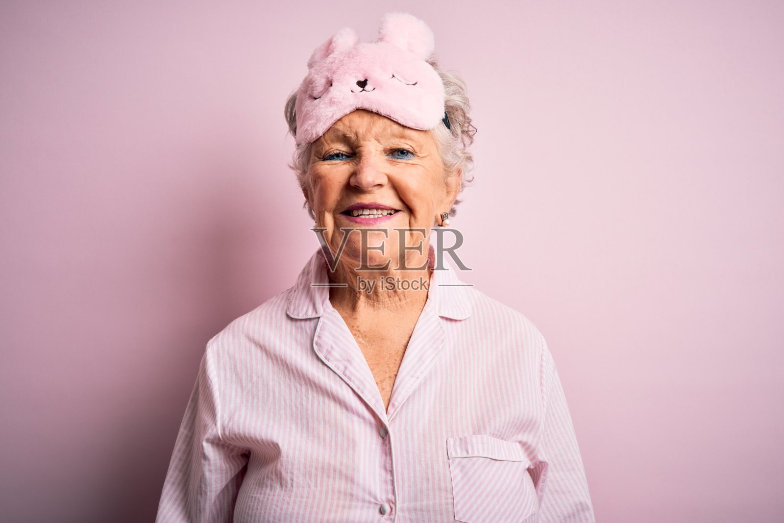 高级美丽的女人戴着睡眠面具和睡衣在孤立的粉红色背景与一个快乐和凉爽的微笑在脸上。幸运的人。照片摄影图片