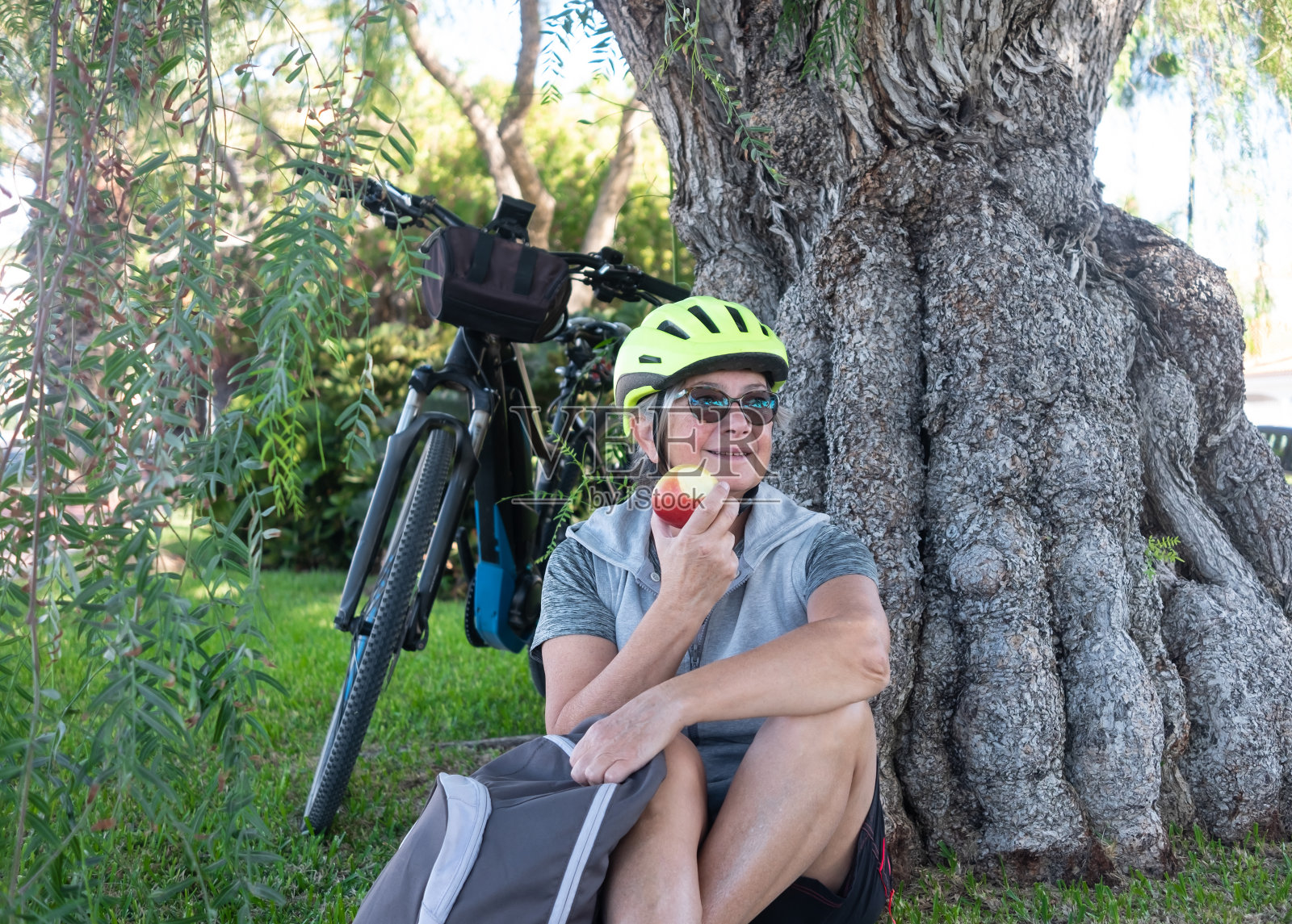 健康的生活方式。有吸引力的资深女子骑自行车与黄色头盔有一个休息在公园附近的一个老和大的原木。吃个红苹果,放松一下。照片摄影图片