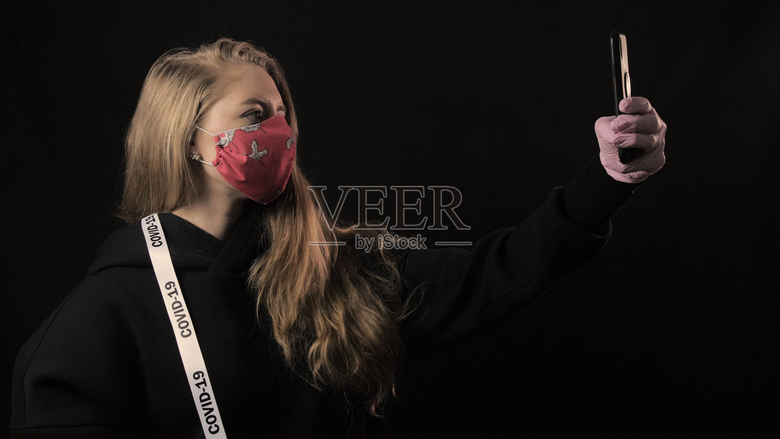 戴着红色口罩,拿着带有“covid 19”字样的白色丝带的女孩用智能手机自拍。黑色背景。医疗保健概念。新冠疫情照片摄影图片