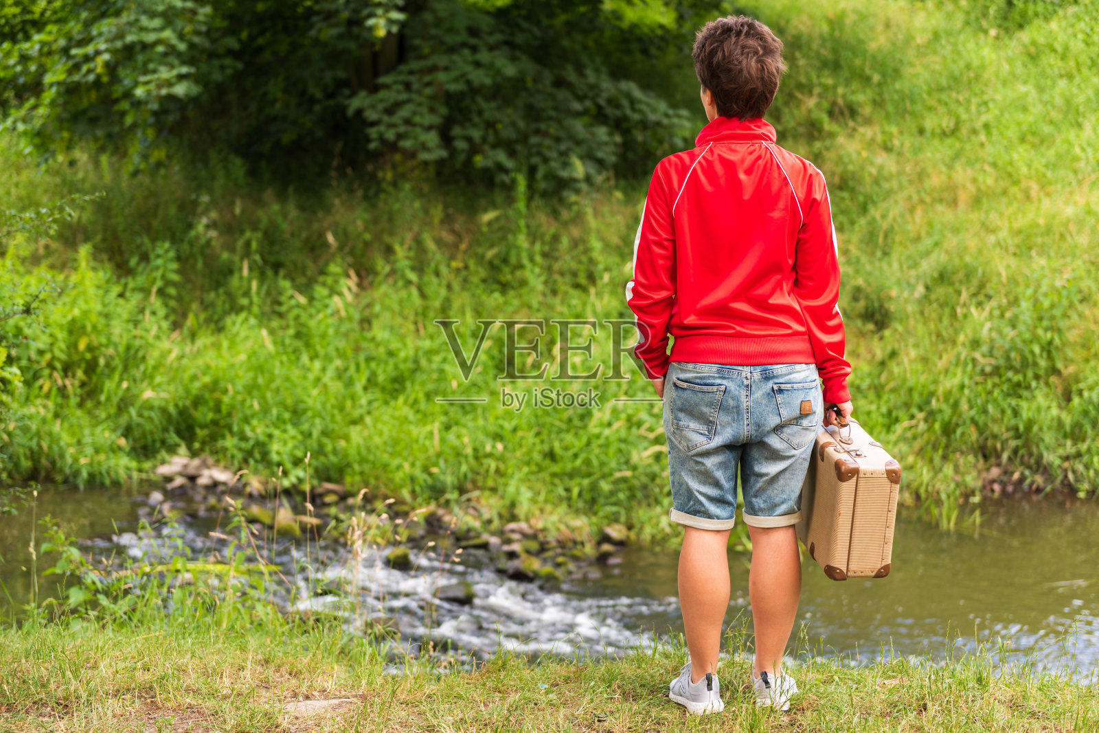 一位拿着复古手提箱的年轻女子坐在河边,背影,本地旅行概念照片摄影图片