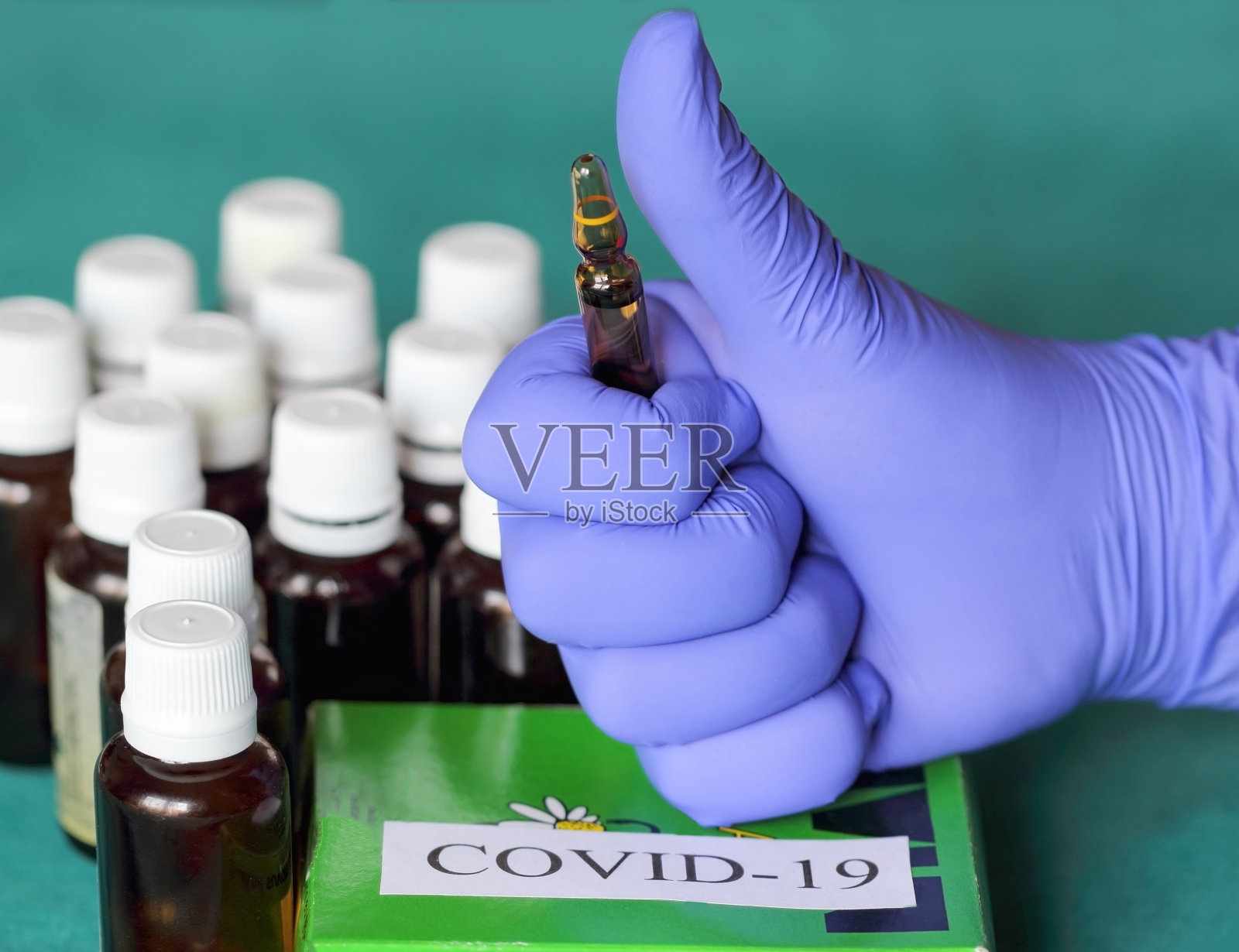 一位戴着蓝色手套的妇女的手倚靠在一个标有Covid -19的盒子上,她举起拇指举着安瓿,像这样。照片摄影图片