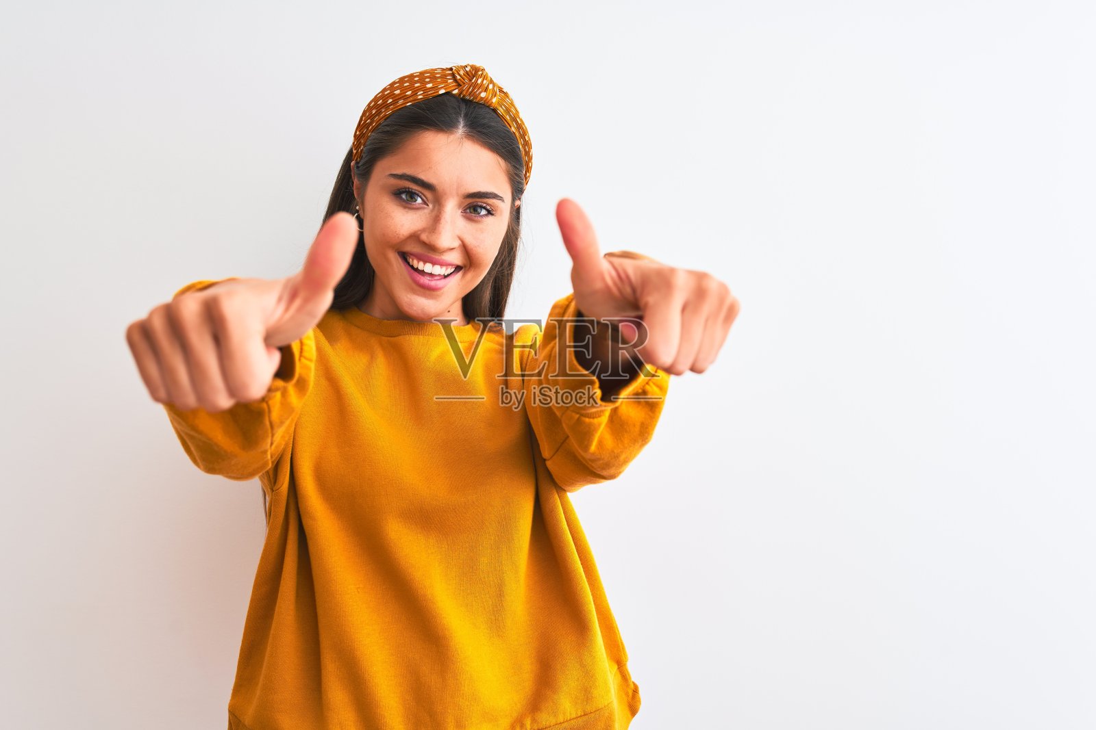 年轻美丽的女人穿着黄色毛衣和冠冕,在孤立的白色背景下表示赞许,做积极的手势,竖起大拇指,微笑,为成功而高兴。赢家的姿态。照片摄影图片