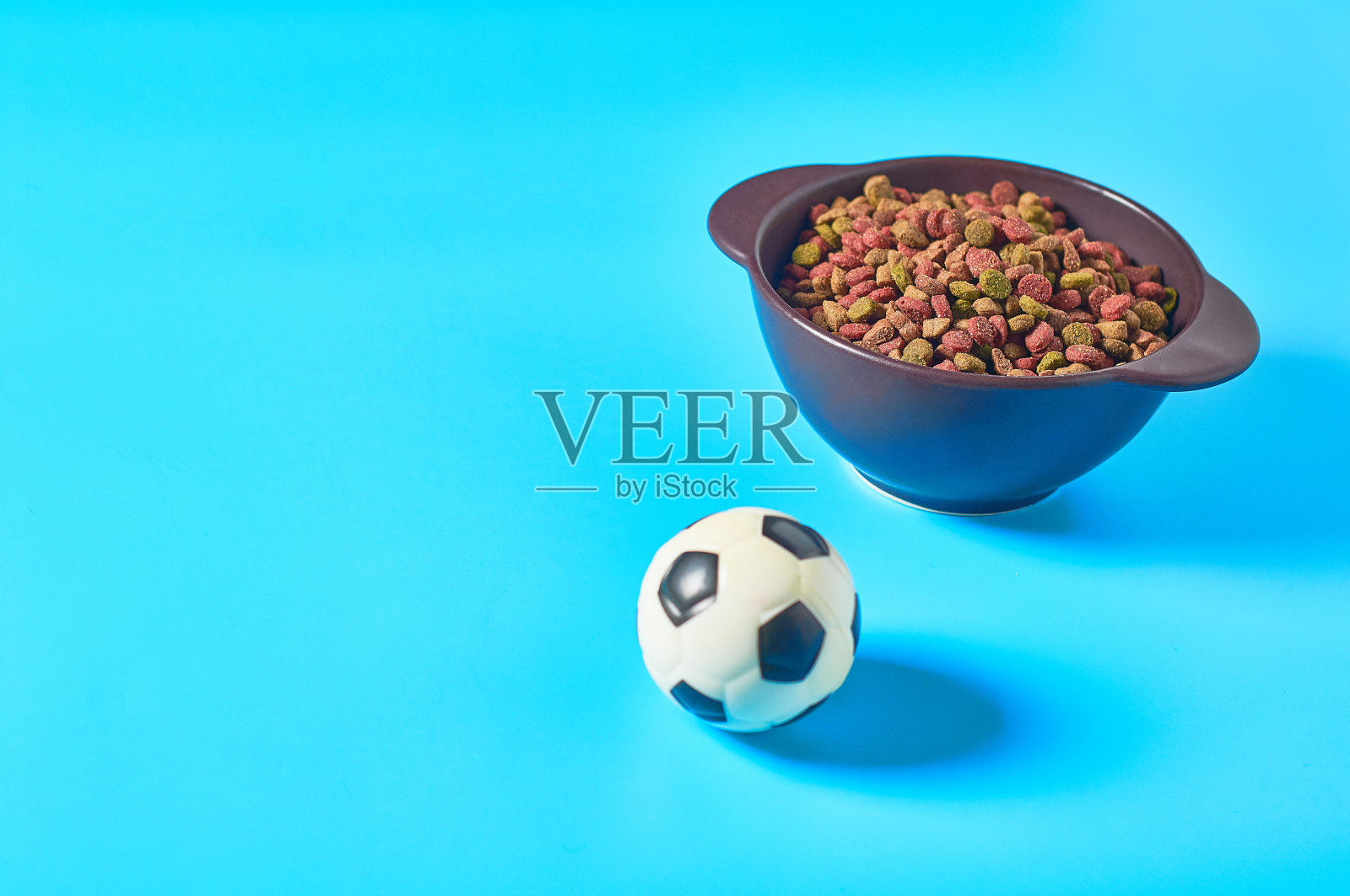 蓝色背景的橡胶球旁边的陶瓷碗里装满了宠物的干粮。本空间照片摄影图片