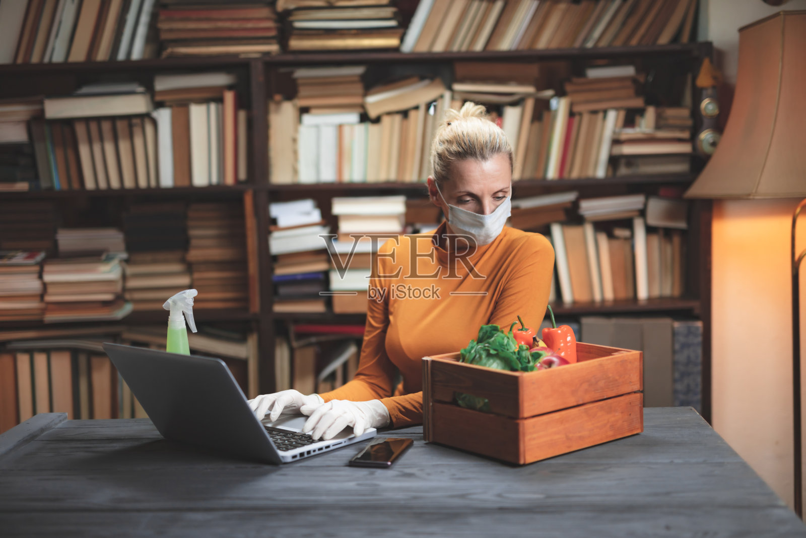 妇女戴着防护口罩,使用笔记本电脑在家,通过互联网订购食品、杂货和供应品。照片摄影图片