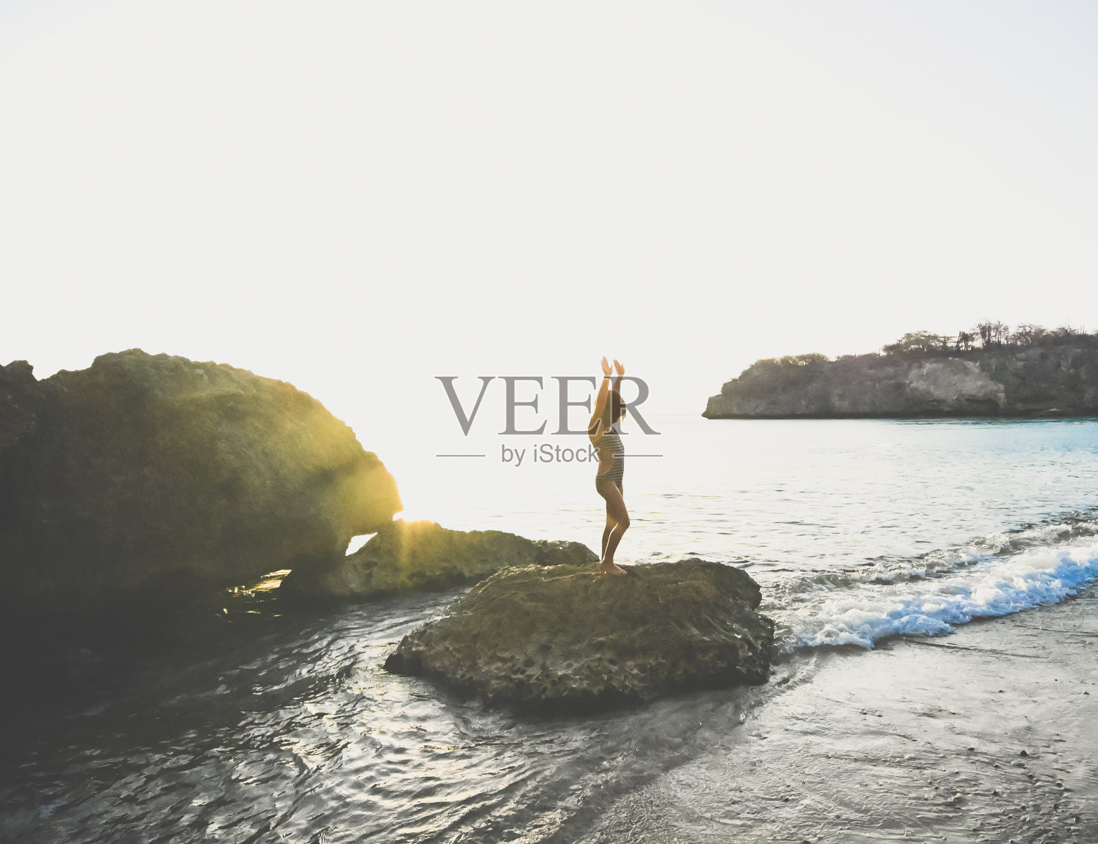 美丽的年轻女孩站在海洋泻湖的一块岩石上,夕阳西下照片摄影图片