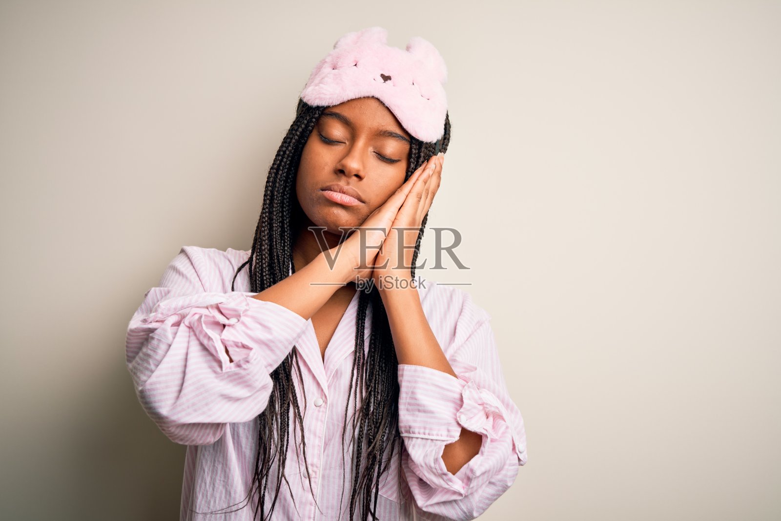 在孤立的背景下,戴着粉红色睡衣和睡眠面罩的年轻非裔美国妇女,睡得很累,做着梦,双手合十,闭着眼睛微笑照片摄影图片