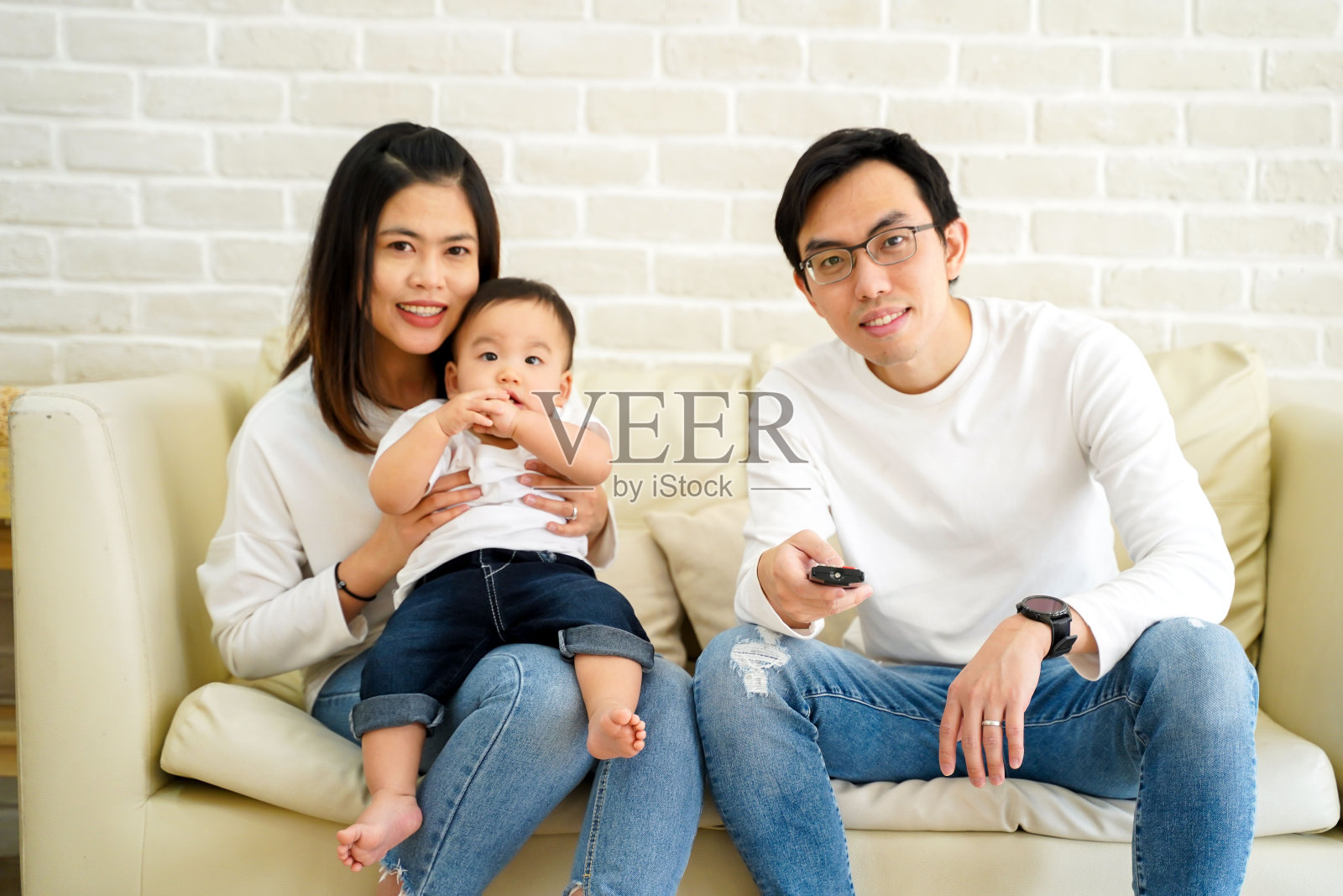 30多岁的幸福亚洲家庭坐在家里的沙发上看电视微笑。父亲手里拿着遥控器。母亲抱着自己的孩子坐在客厅的沙发上。照片摄影图片