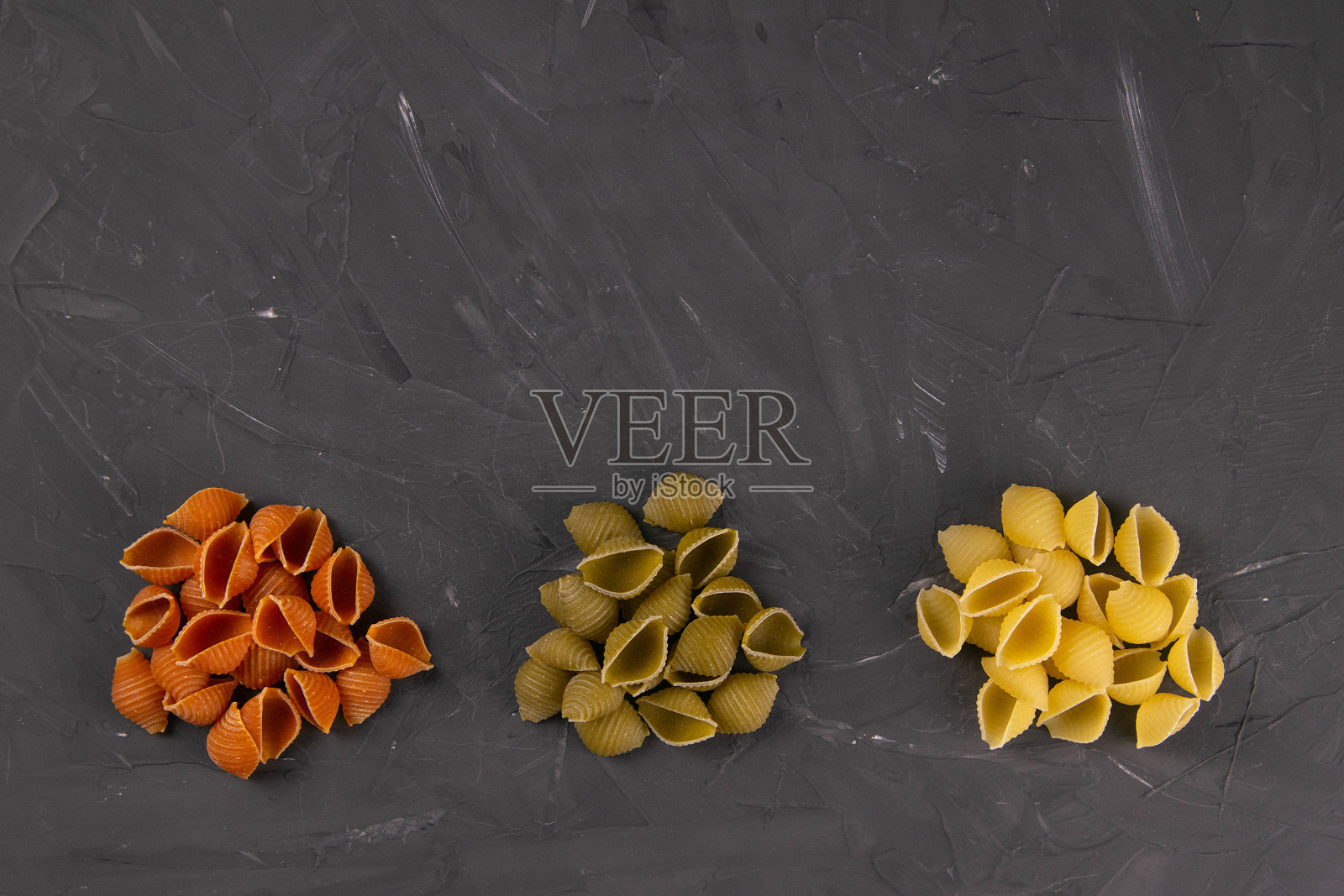 在黑板背景上的干贝壳意面俯视图。三个圆形的幻灯片,上面铺着红色、绿色和黄色的贝壳意面照片摄影图片