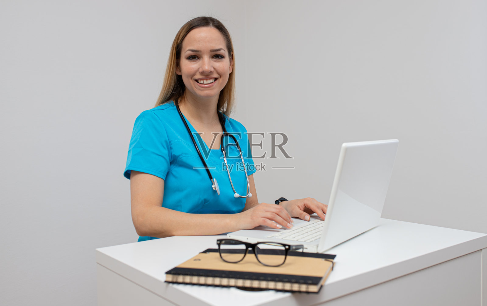 自信的护士坐在办公室里。自信的年轻女医生。实习护士。年轻漂亮的女人在白色衣服与听诊器摆姿势和微笑。照片摄影图片