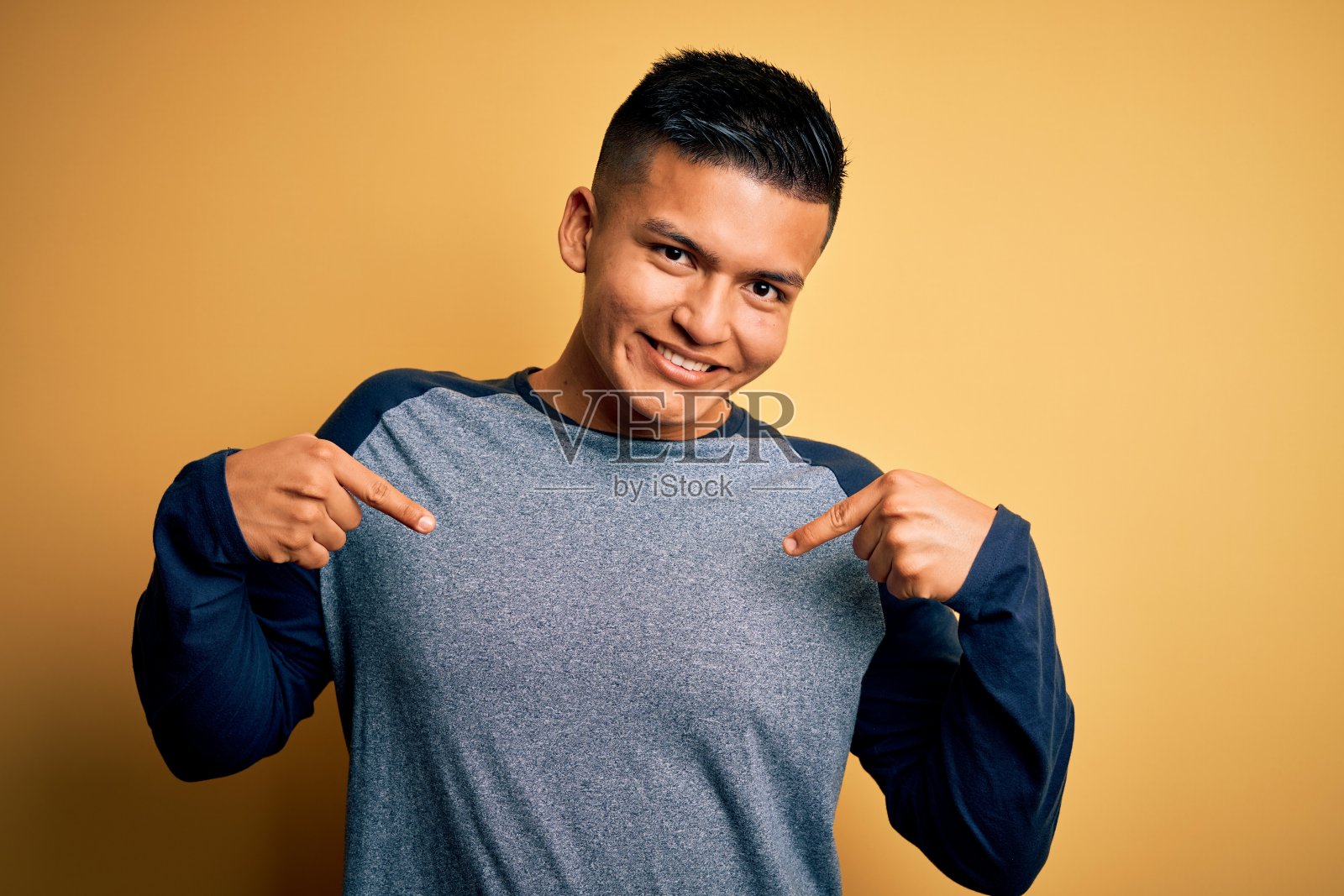 年轻英俊的拉丁男子穿着休闲t恤站在黄色的背景看起来自信,脸上带着微笑,用手指指着自己骄傲和幸福。照片摄影图片