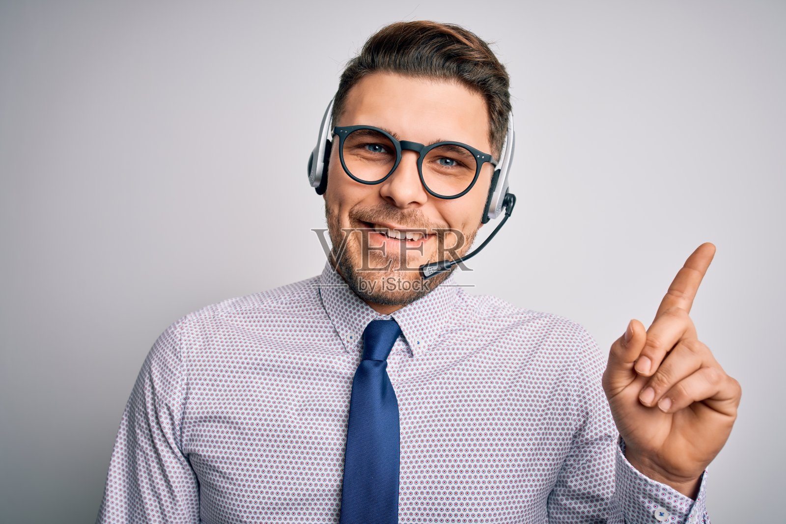 年轻的呼叫中心话务员戴着蓝眼睛和耳机,脸上带着大大的笑容,用手和手指指着一边看着摄像头。照片摄影图片