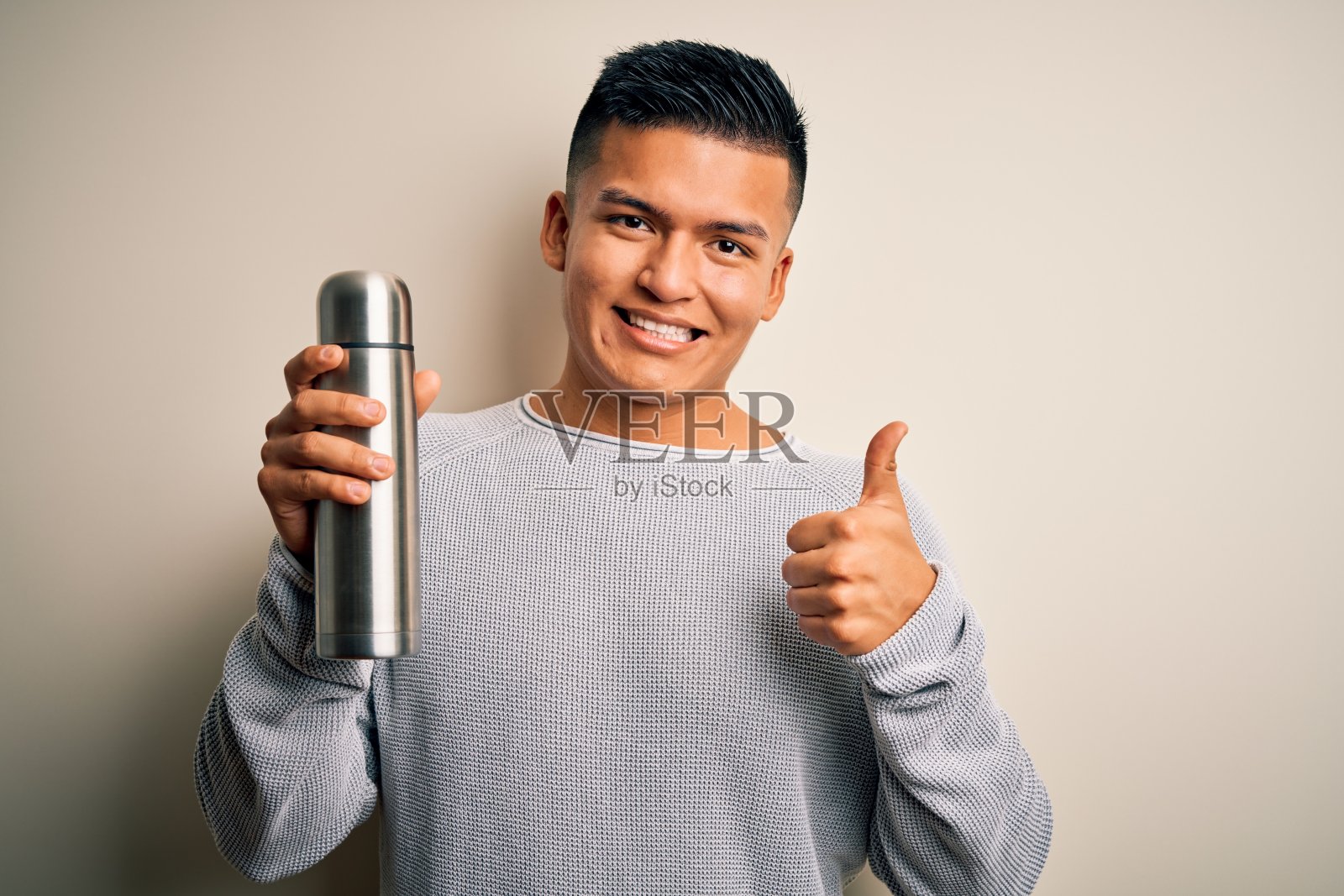 年轻英俊的拉丁男子拿着热水与水在孤立的白色背景愉快地与大微笑做ok的手势,拇指向上的手指,优秀的手势照片摄影图片