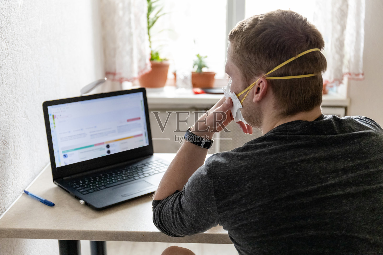 一名戴着医用防护口罩的年轻人在家里用笔记本电脑工作,在大流行期间进行家庭隔离照片摄影图片