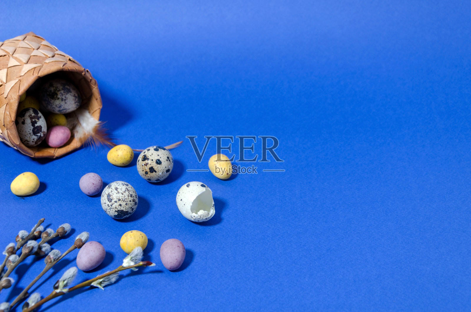 鹌鹑蛋和彩色的迷你果冻豆蛋躺在蓝色时尚的背景。旁边放着一个小柳条篮子,里面有鸡蛋和嫩柳条。复活节属性,可爱的春天背景。水平的形象照片摄影图片
