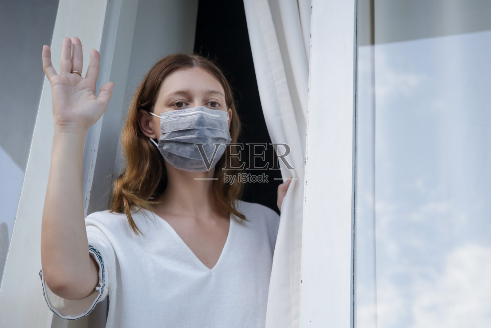 年轻女子戴着一次性医用口罩透过窗户看外面。无聊,一个人在家。冠状病毒爆发和自我隔离概念。照片摄影图片