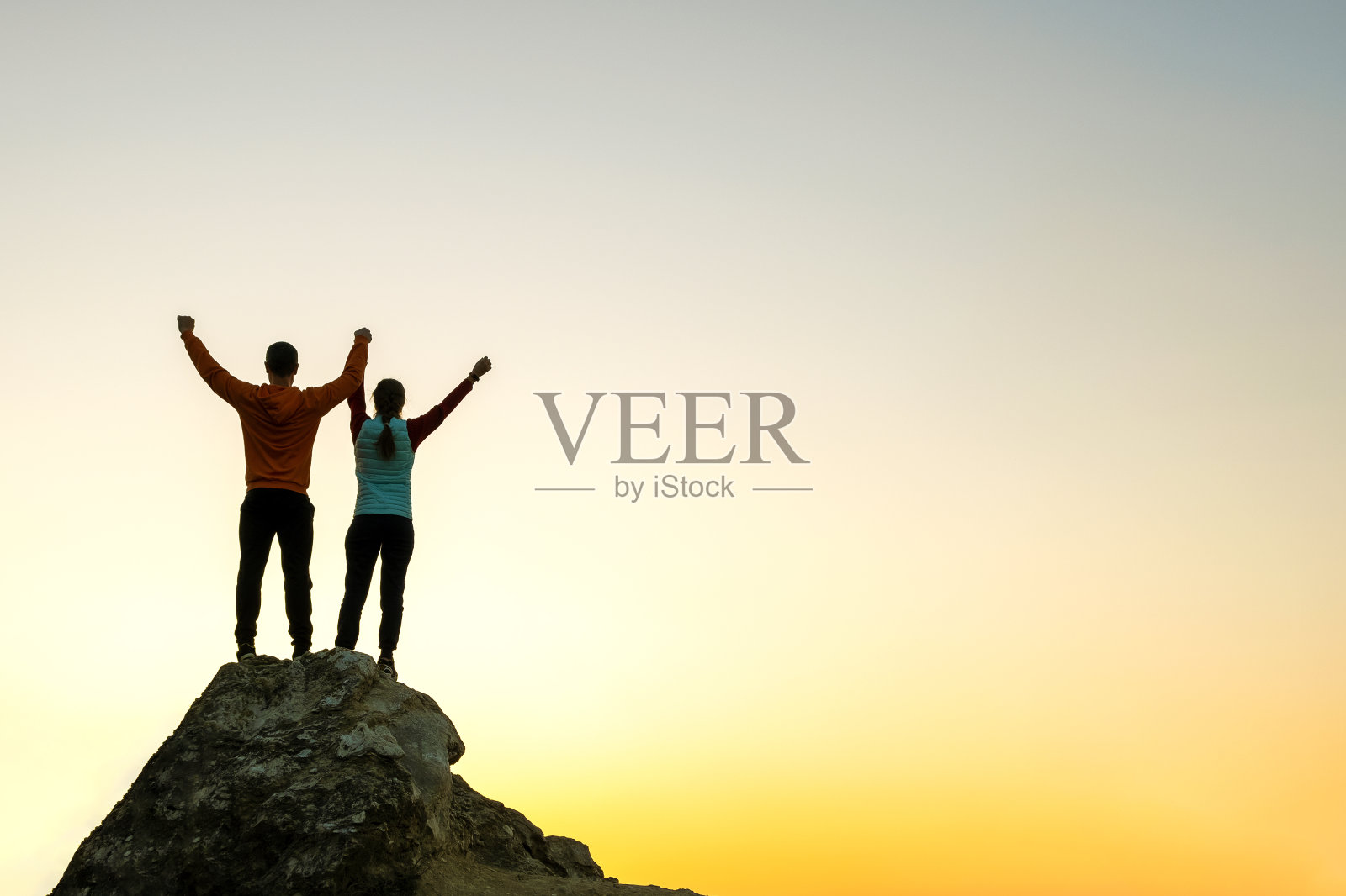 日落时分,一男一女徒步旅行者站在山上的一块大石头上。傍晚时分,一对夫妇在高高的岩石上举起双手。旅游、旅游和健康的生活理念。照片摄影图片