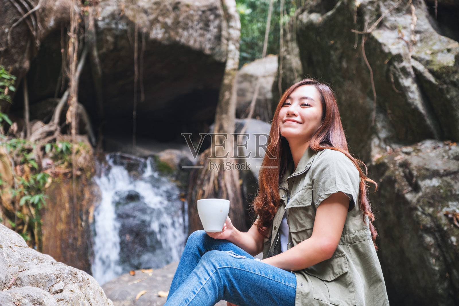 坐在瀑布前的岩石上喝咖啡的女人照片摄影图片
