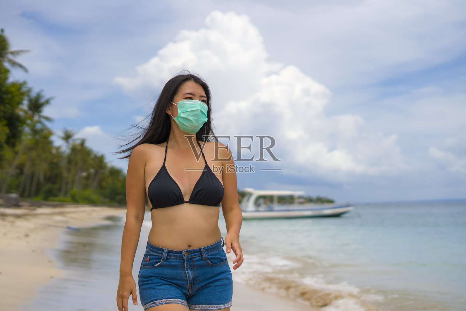 在公共场所使用医用口罩——年轻貌美的华裔女子穿着比基尼和防护口罩在海滩度假,以预防病毒感染照片摄影图片