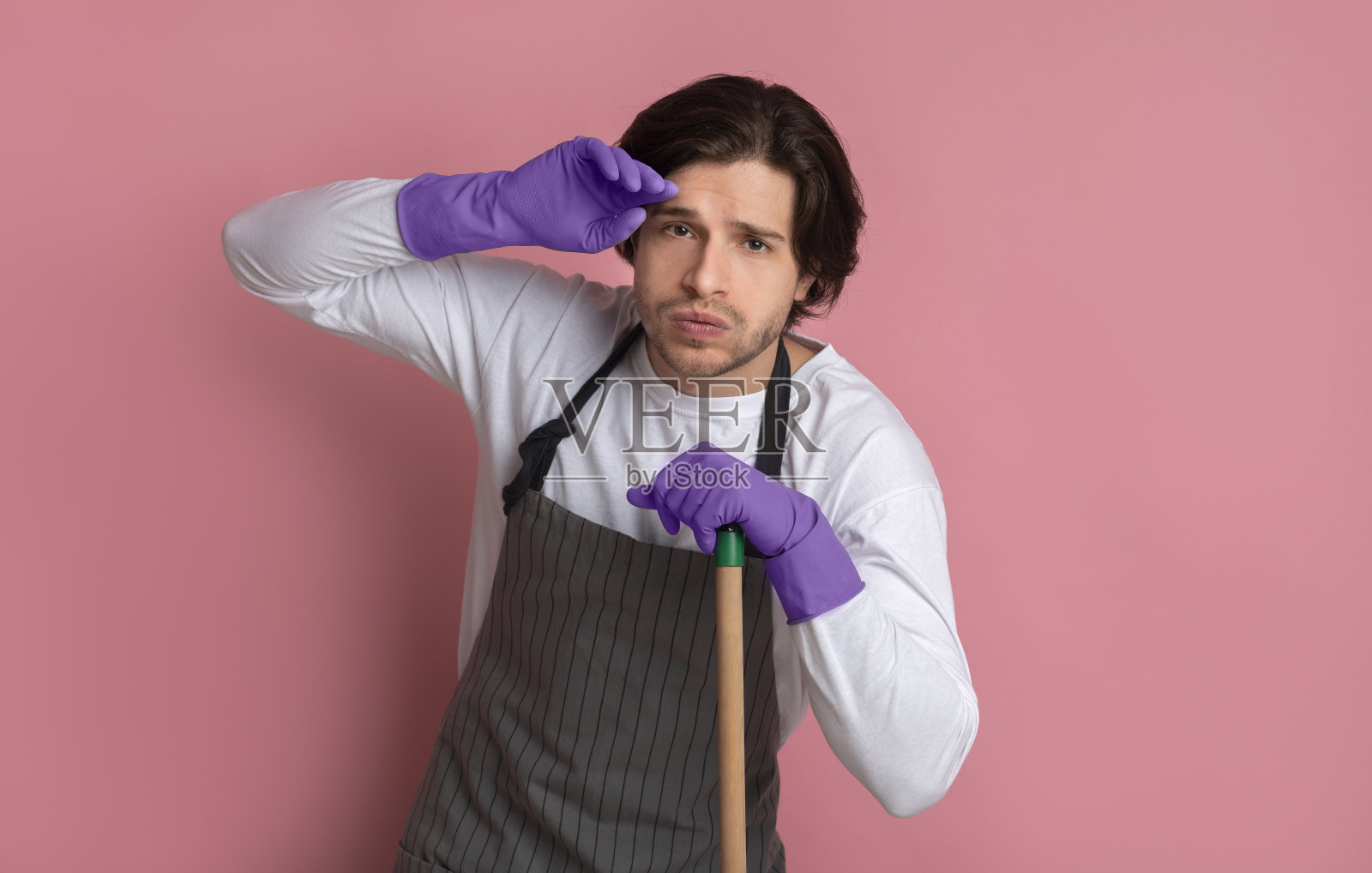 清洁后疲惫地靠在拖把上的年轻男人,粉色背景照片摄影图片