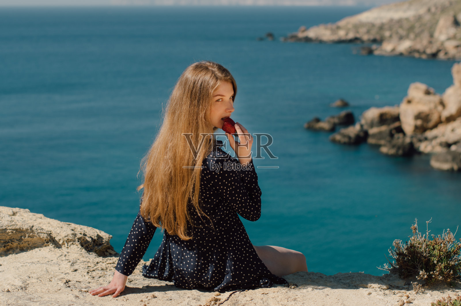 美丽微笑的金发女孩,吃着草莓,坐在山的边缘,看着大海照片摄影图片
