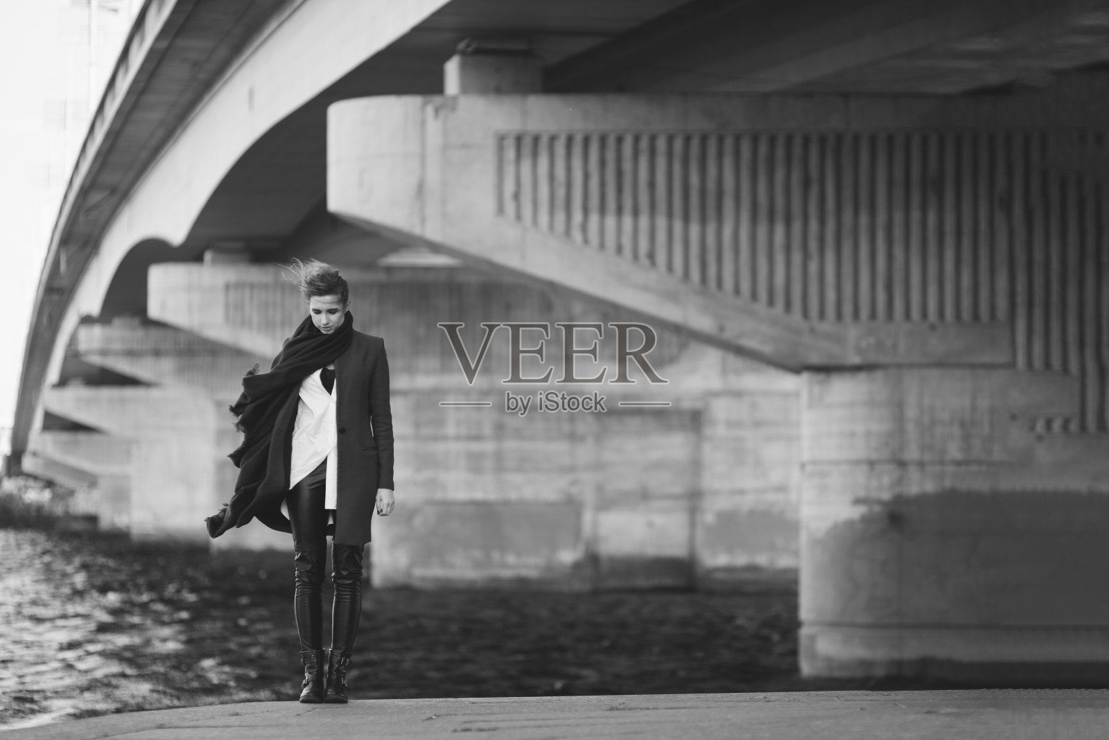 穿着大衣和围巾的美丽孤独的女人在河边的桥下随风飘动。孤独的概念。黑白艺术照片。软的焦点。照片摄影图片