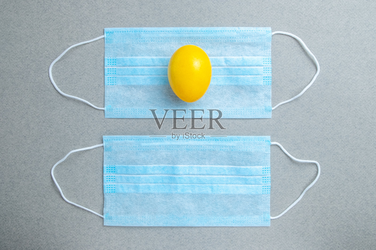 一个黄色的复活节彩蛋放在一个蓝色的医用面具上,背景是灰色的。照片摄影图片