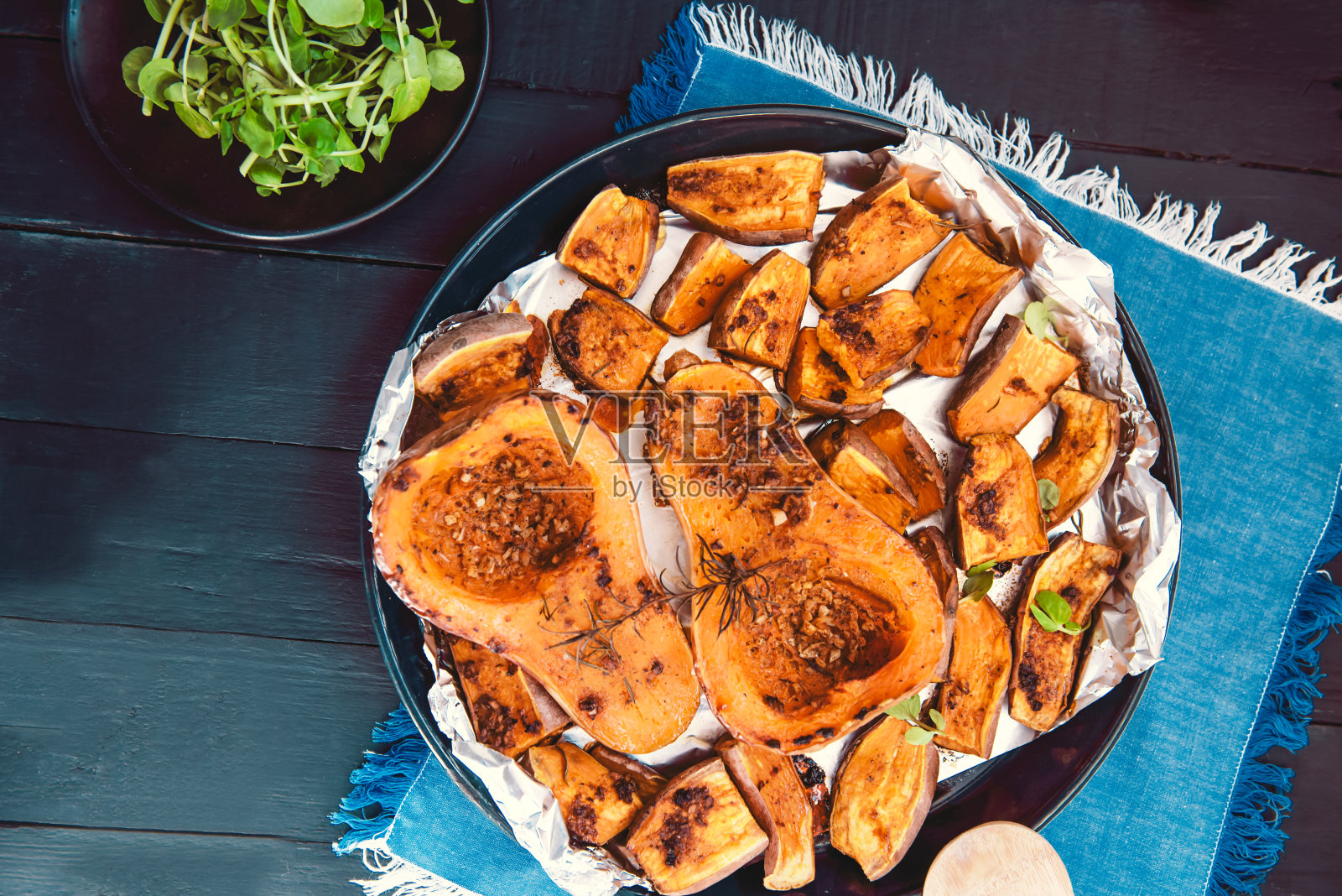 俯视图烤烤烤整个橙色南瓜、冬南瓜和带香草的甘薯片放在烤盘上,蓝色的餐巾和黑色的木桌。素食和纯素饮食。照片摄影图片