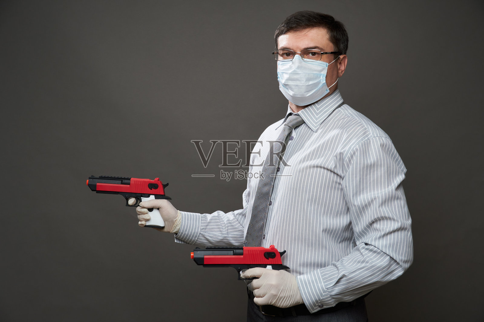 一名男子扮成商人,在灰色背景的演播室里摆姿势,戴着医用口罩和防护手套、枪、眼镜、衬衫和领带——概念的检疫和防病毒防护照片摄影图片