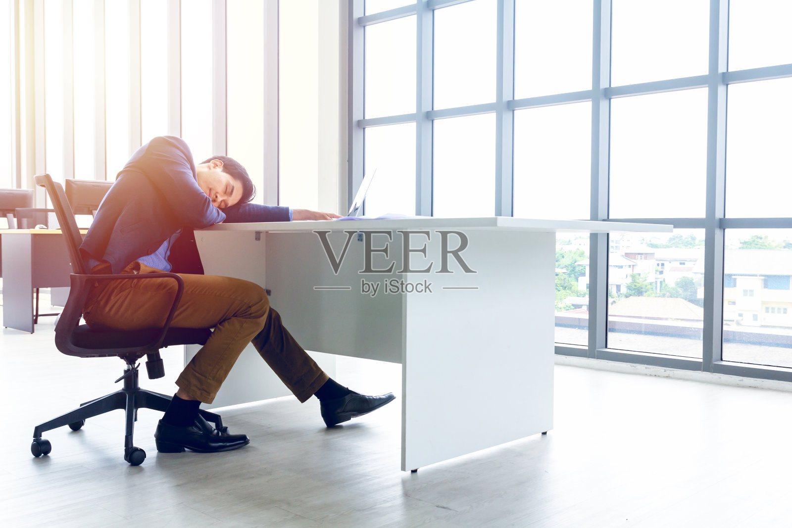 一个年轻的商人坐在一间现代化的办公室里。他感到困倦,因为工作太辛苦了,疲惫不堪。在他的桌子上有一台电脑,笔记本电脑,写字板,笔,纸图形。照片摄影图片