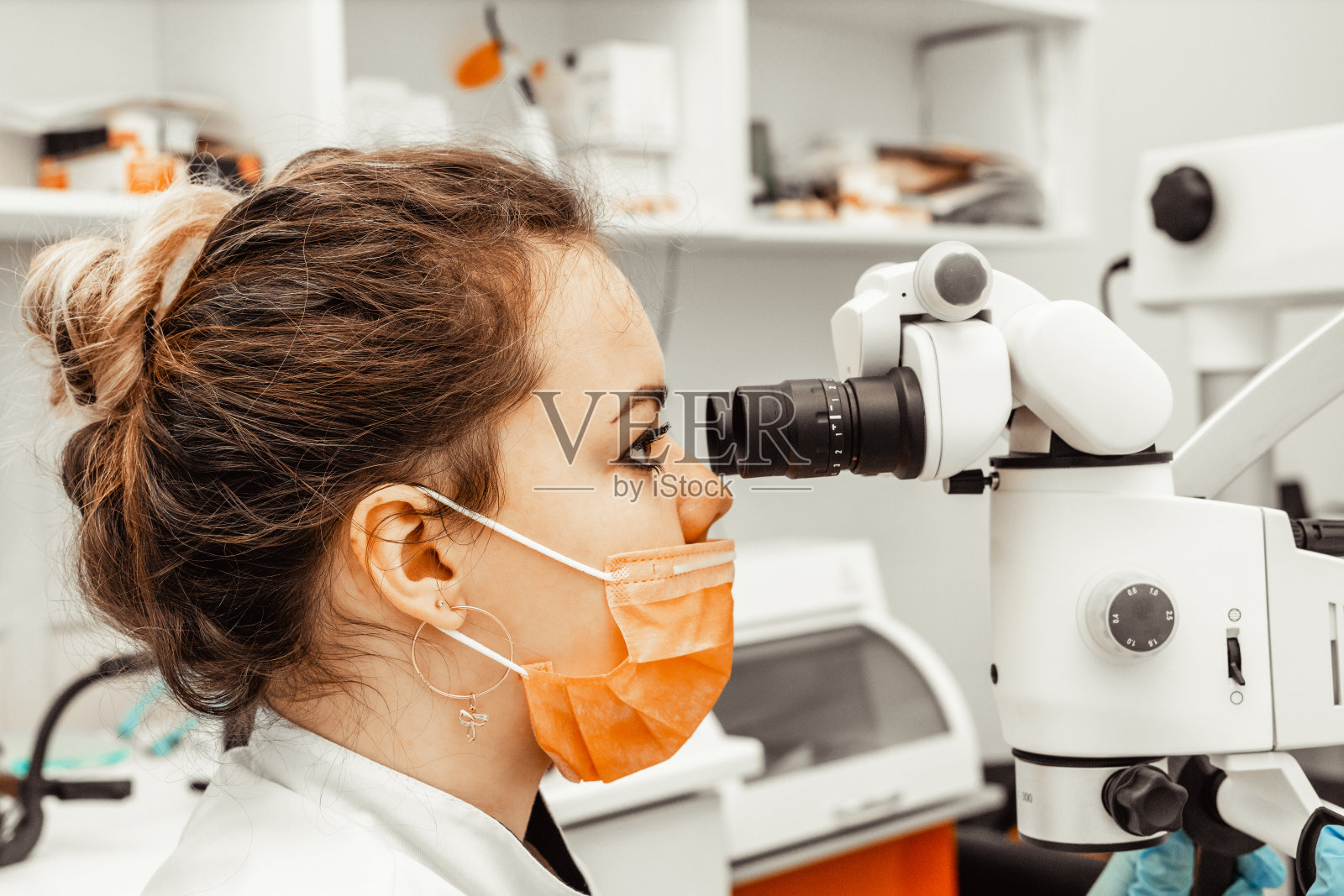 一位年轻的女牙医在牙科诊所里透过专业显微镜看牙医。戴一次性医用口罩和帽子的医生。牙科的先进设备照片摄影图片