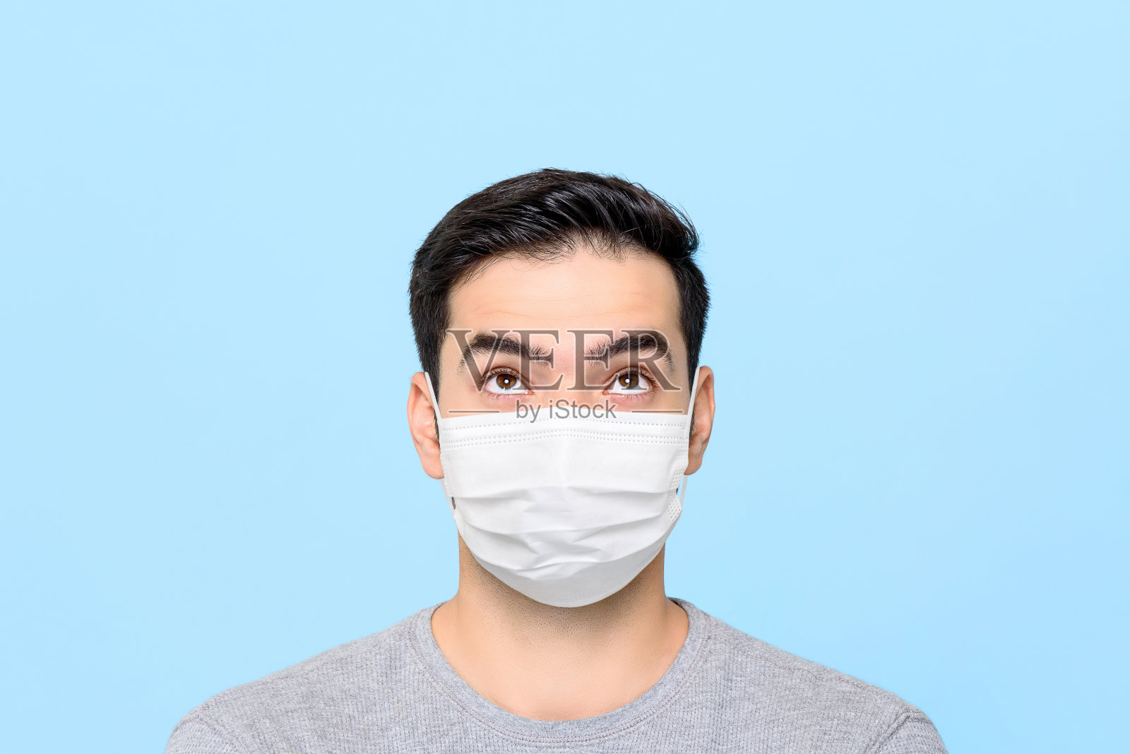 年轻男子戴着医用口罩思考着,仰望着上方孤立在浅蓝色演播室背景下的空旷空间照片摄影图片