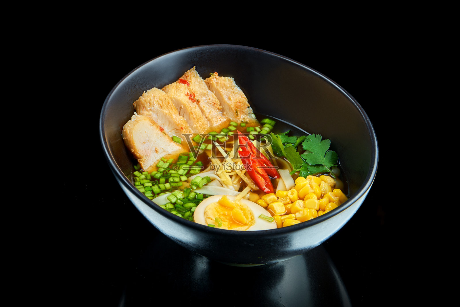 传统的亚洲拉面汤,用洋葱,鸡蛋,猪肉,玉米,欧芹和辣椒放在一个黑色的碗里,在黑色的背景下反射照片摄影图片