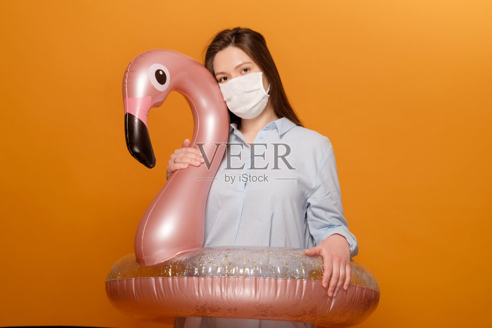 办公室职员年轻美丽的女人在橙色黄色的背景在保护医疗面具拥抱游泳圈粉红色火烈鸟在抑郁症,流行病,假期取消,隔离的女人照片摄影图片