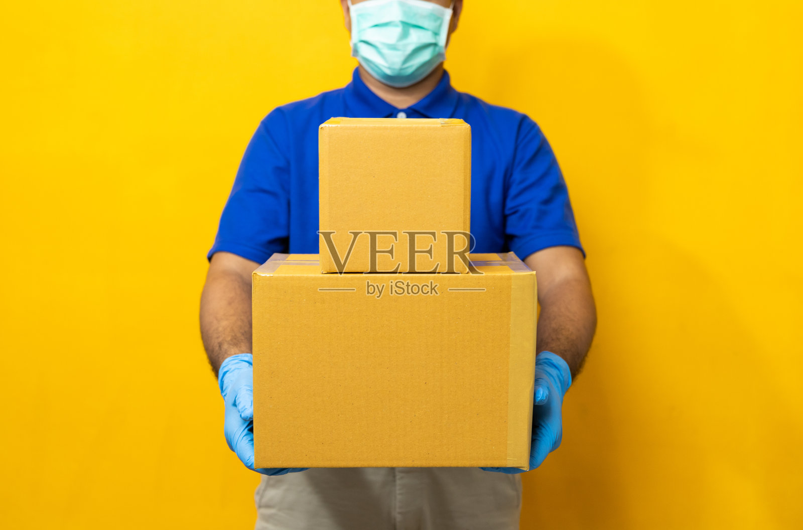 快递员身穿蓝色制服,戴着橡胶手套和口罩,拿着黄色背景的包裹纸箱。照片摄影图片