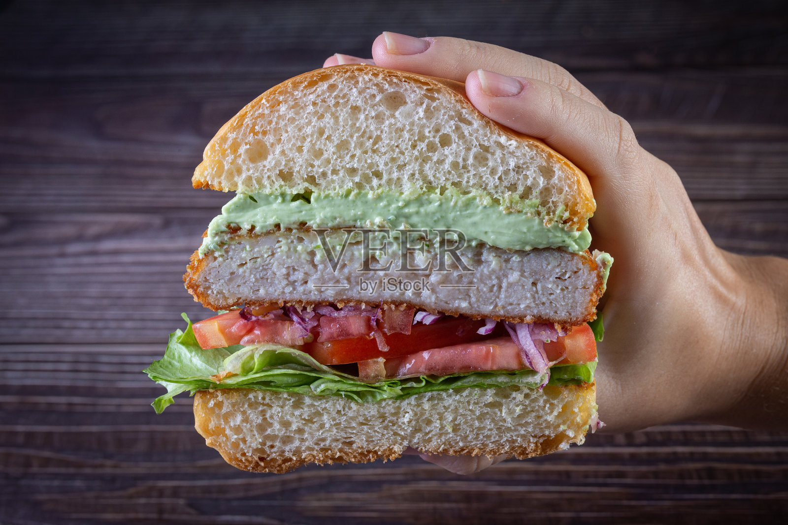 手拿鸡肉汉堡与生菜,番茄,紫洋葱和手工蛋黄酱在木材背景。美味。照片摄影图片