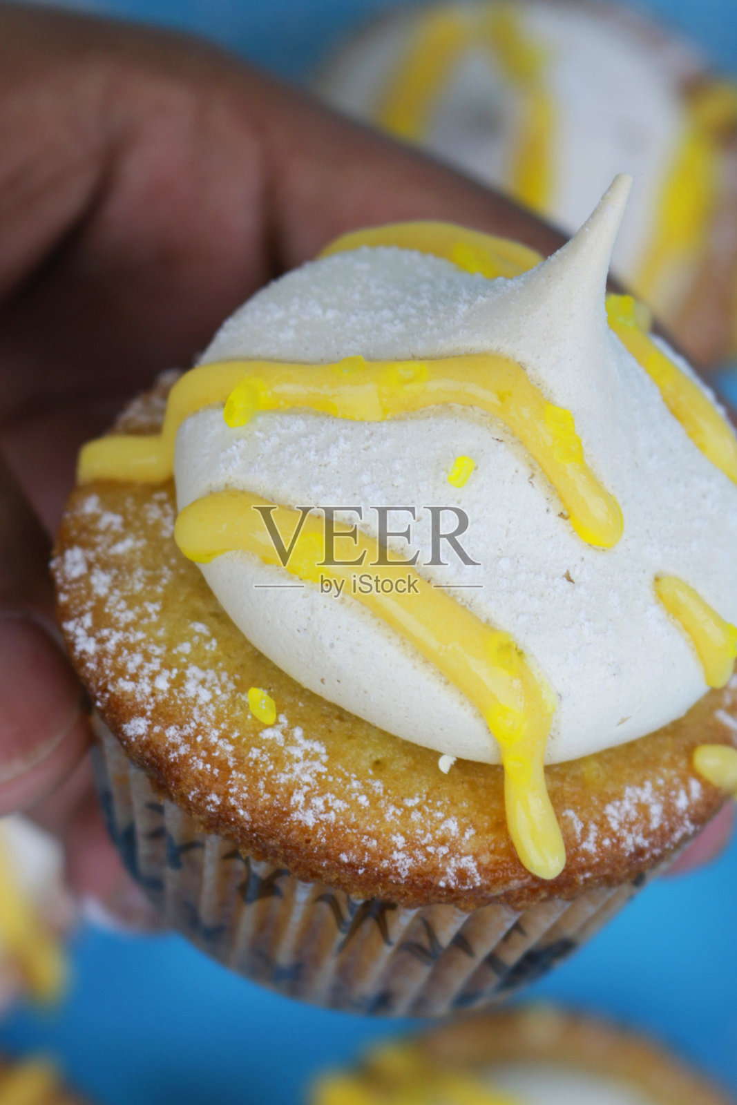 这是自制柠檬纸杯蛋糕的特写,上面撒着迷你蛋白酥皮,淋上柠檬凝乳,撒上糖霜,拿在手里,蓝色的背景照片摄影图片