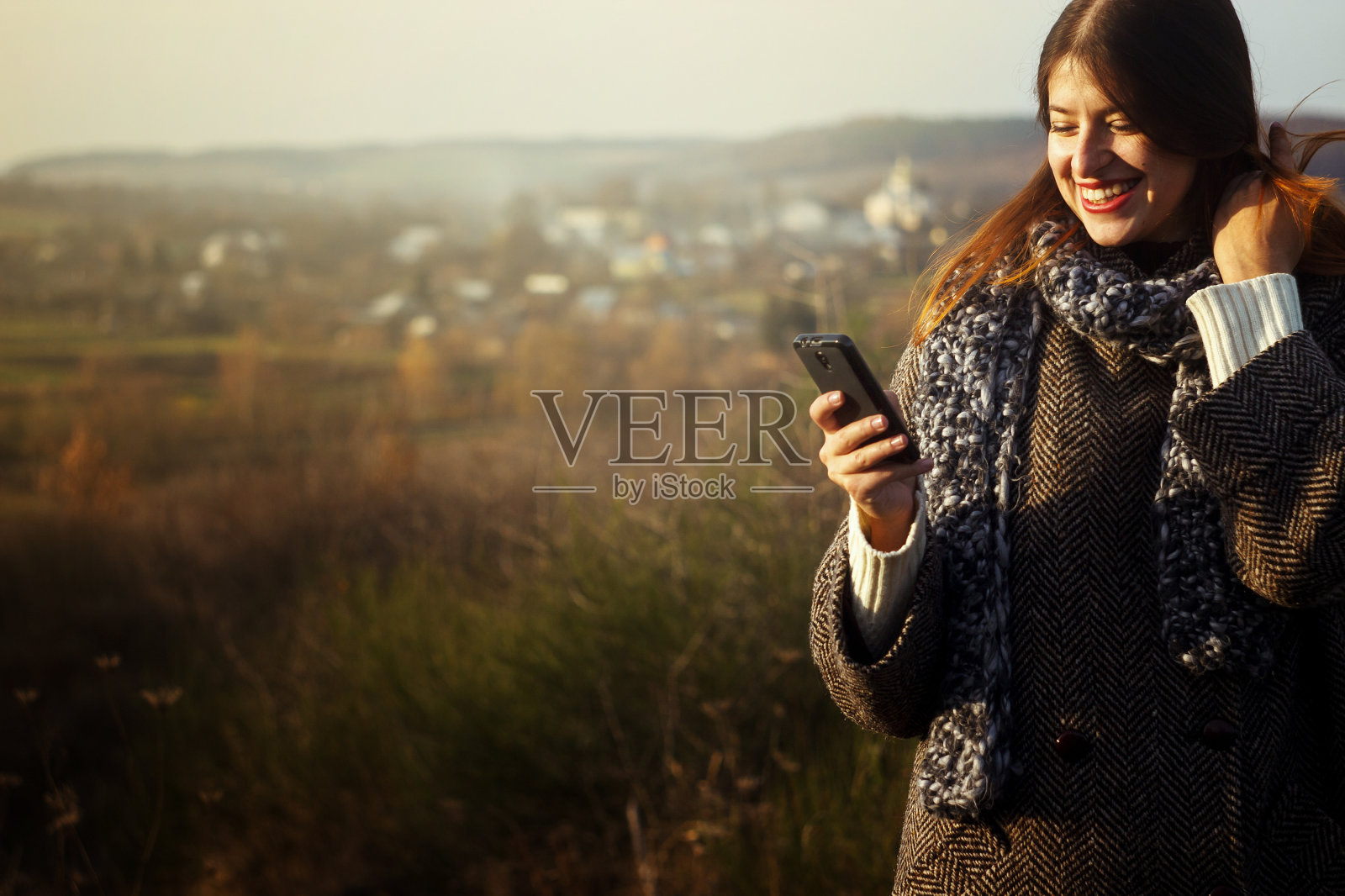 华丽时尚美丽快乐的黑发女孩看着电话和微笑在一个阳光明媚的秋天的自然背景照片摄影图片