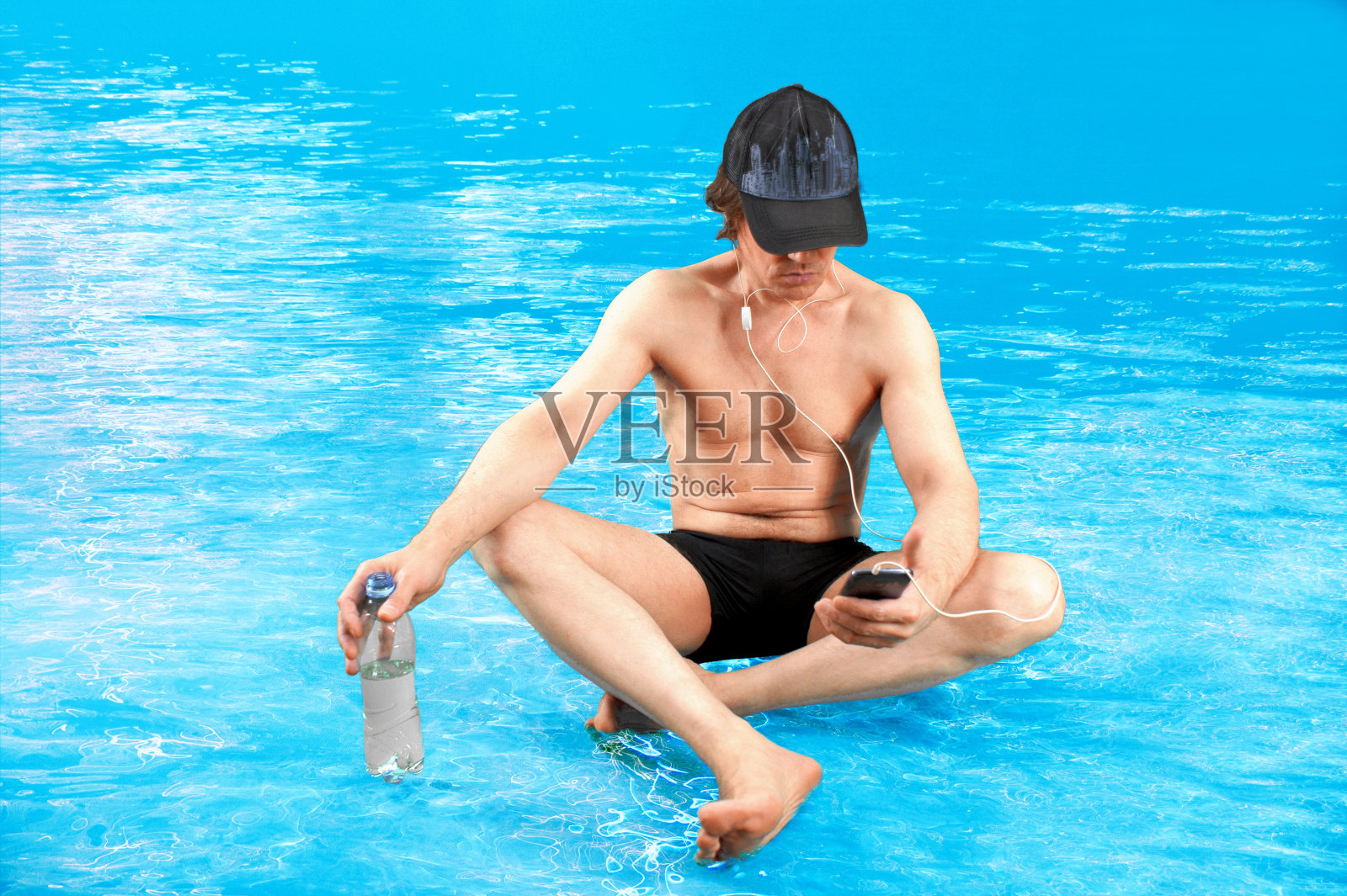 有魅力的运动员盘腿坐在水里,晒日光浴,用智能手机听音乐,用瓶子喝饮料。照片摄影图片