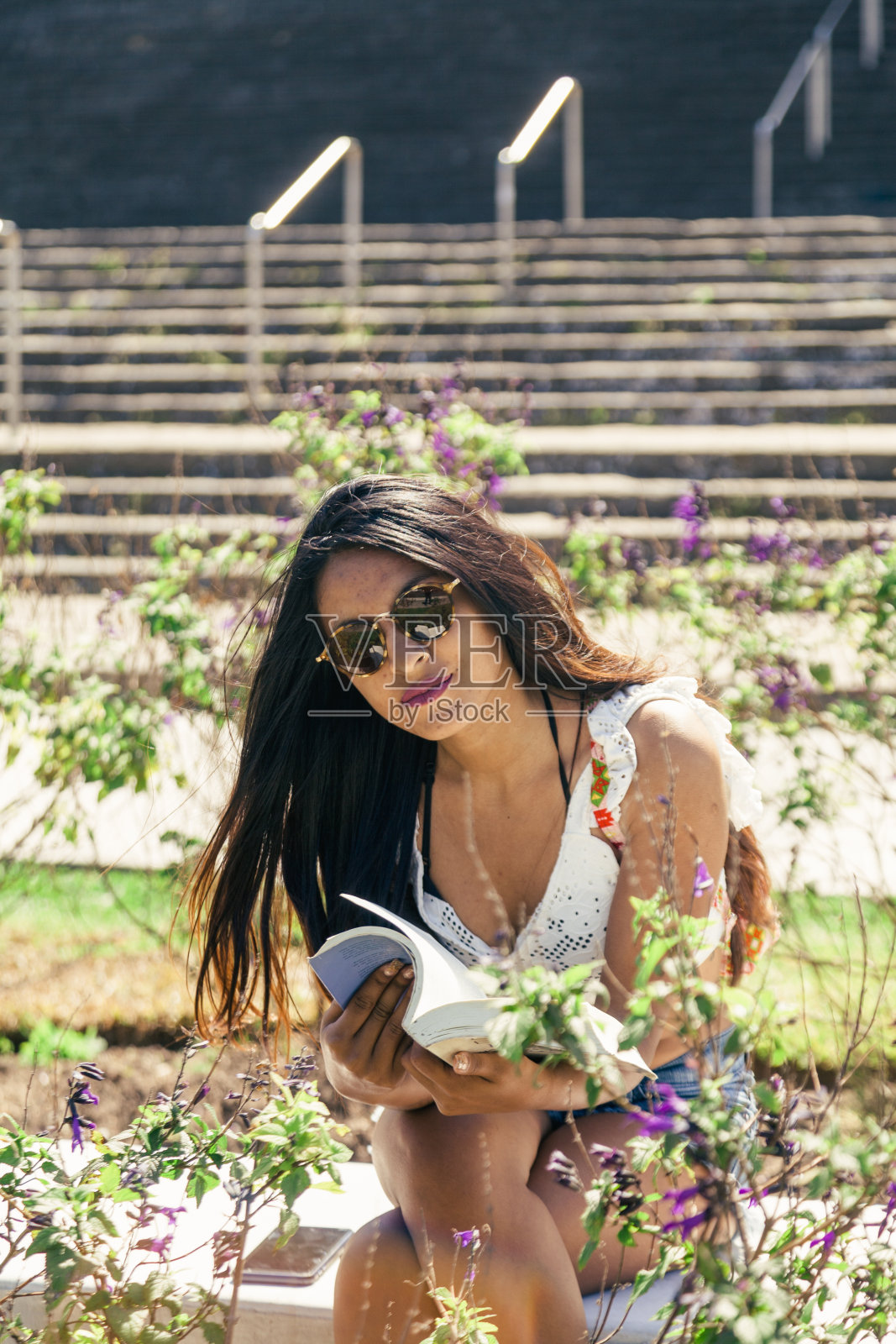 女孩戴着太阳镜,穿着夏装,坐在花园里的长凳上,拿着一本打开的书照片摄影图片