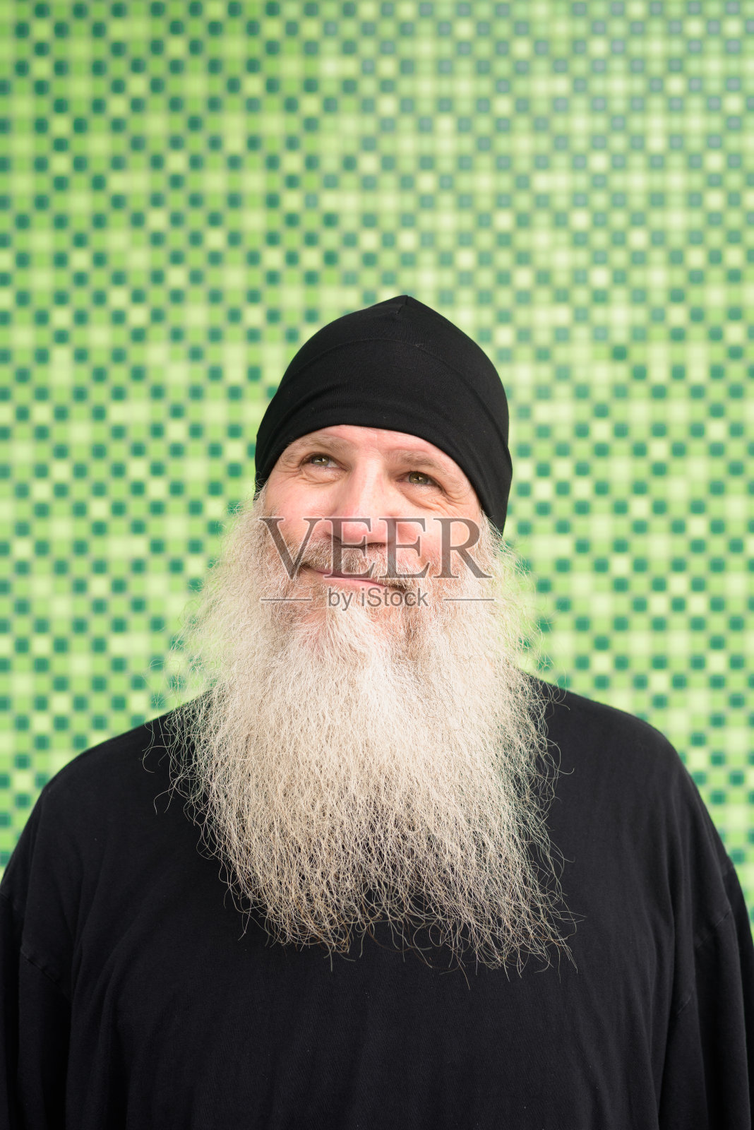 蓄着大胡子、戴着无檐便帽、对着绿墙思考的成熟嬉皮士照片摄影图片