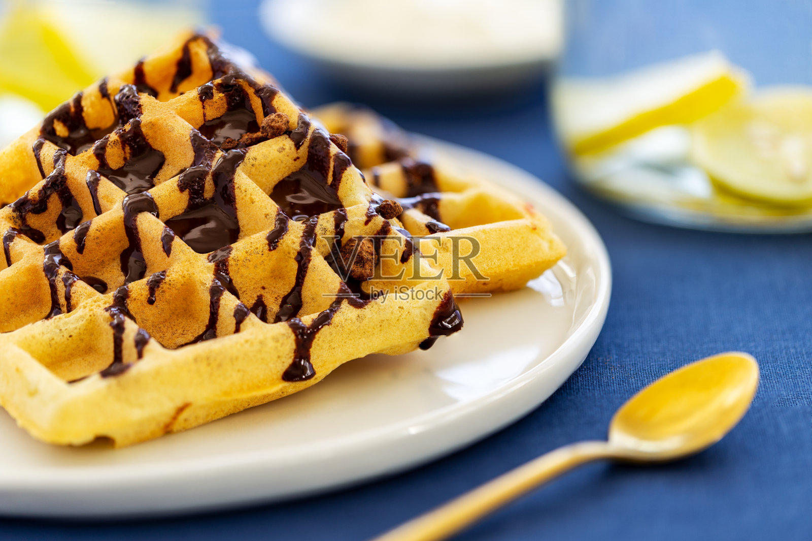 特写比利时华夫饼,上面淋上巧克力酱,放在盘子里,旁边是一个金色的勺子和金色的钳子,背景是蓝色的布。照片摄影图片